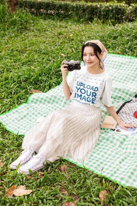 T-Shirt Mockup Featuring a Young Woman Holding a Vintage Camera on a Picnic Blanket in a Park