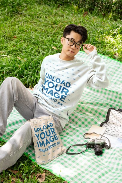 Sweatshirt Mockup Featuring a Young Man Adjusting His Glasses on a Picnic Blanket Outdoors