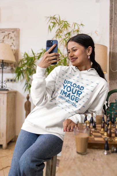 Hoodie Mockup Featuring a Smiling Young Woman Taking a Selfie Indoors Next to a Chessboard