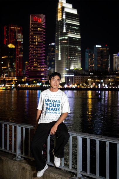 T-Shirt Mockup Featuring a Young Man Sitting on a Railing With a City Skyline at Night m52860
