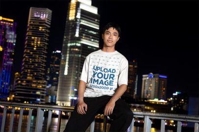 T-Shirt Mockup Featuring a Young Man Sitting on a Railing With a City Skyline at Night