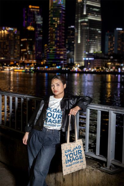 Tote Bag and T-Shirt Mockup of a Woman in a Leather Jacket Standing by a Riverwalk at Night