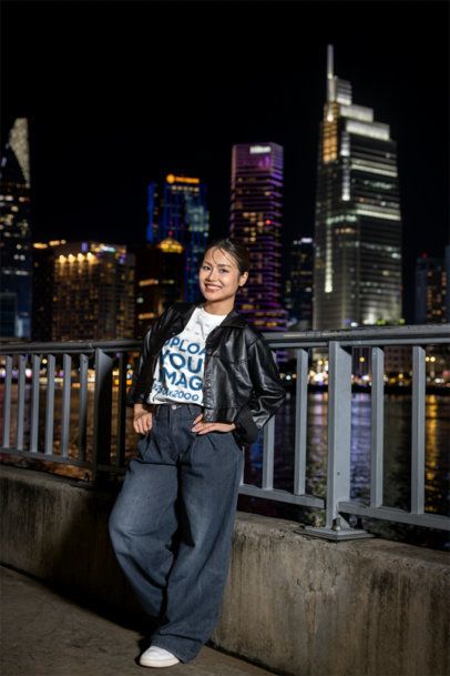 T-Shirt Mockup Featuring a Smiling Young Woman Standing by a Railing With a City Skyline at Night