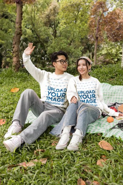 Couples Sweatshirt Mockup Featuring a Young Man Waving While Sitting Next to a Smiling Woman on a Picnic Blanket in a Park m57810