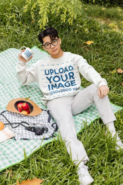 Sweatshirt Mockup Featuring a Young Man Holding a Mug on a Picnic Blanket in a Park m57779