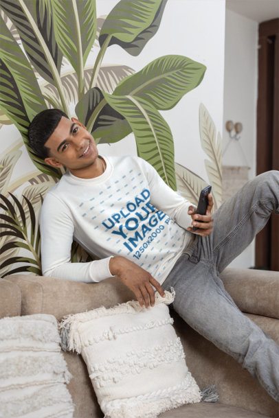 Sweatshirt Mockup Featuring a Smiling Young Man Holding a Smartphone on a Couch