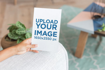 Mockup of a Hand Holding a Flyer on an Armchair