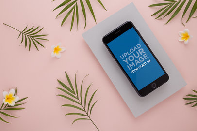 Space Gray iPhone 8+ Mockup Placed on a Rectangular Gray Cardboard over a Pink Surface with White Flowers and Leaves