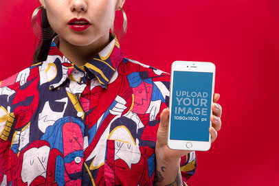 Mockup of a Woman with Hoop Earrings Wearing a Colorful Shirt Showing a Silver iPhone 8 Against a Red Background 21961