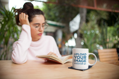 Mockup of a Coffee Mug on a Table in Front of a Woman Reading a Book