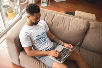 T-Shirt Mockup of a Man with an Arm Tattoo Using his Laptop at Home