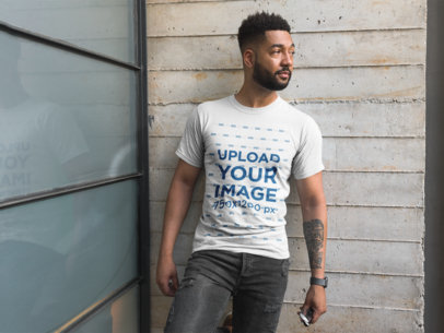 Front View T-Shirt Mockup Featuring a Man with a Beard Against a Concrete Wall and a Window