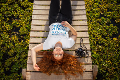 Mockup of a Red-Haired Woman Wearing a T-Shirt while Resting on a Wooden Bridge