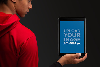 Mockup of a Man with a Red Hoodie Showing an iPad Against a Solid Backdrop 22639