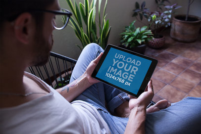 Mockup of a Man with Tattoos Holding an iPad in Landscape Position Sitting on an Acapulco Chair 22816