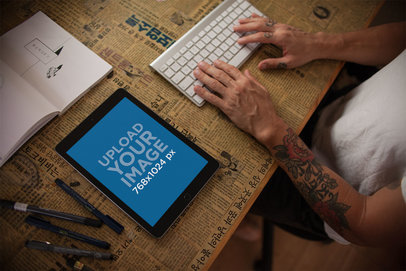 Mockup of an iPad in Portrait Position Lying on a Table Next to a Tattooed Man Using a Keyboard 22832