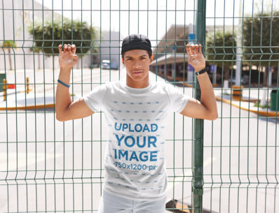 Tshirt Mockup of a Man with a Hat Leaning Against a Fence