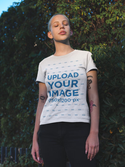 T-Shirt Mockup of a Beautiful Young Woman with a Buzz Cut Haircut Standing in Front of a Hedge