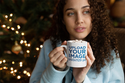 Mug Mockup of a Woman with Curly Hair Drinking Hot Cocoa in Front of a Christmas Tree