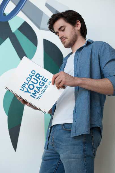 Mockup of a Man Holding a School Notebook by a Decorated Wall 24163