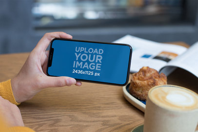 Mockup of an iPhone XS Being Held on Landscape Position Over a Cafe Table 25372
