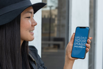Mockup of a Smiling Girl with a Hat Holding Her iPhone XS Max in an Urban Scenario 25771