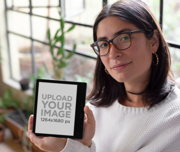 Mockup of a Woman with Glasses Holding a Kindle Oasis by a Window