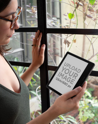 Mockup of a Woman with Glasses Reading from Her Kindle Oasis by Some Plants 26071