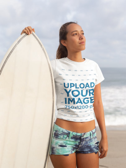 Tee Mockup of a Blonde Woman Standing at the Beach Next to Her Surfboard