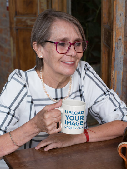 Mockup of an Elderly Woman Holding an 11 Oz Coffee Mug at a Table