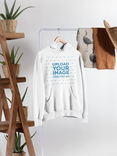Pullover Hoodie Mockup Hanging From a Rack Next to Plant Pots