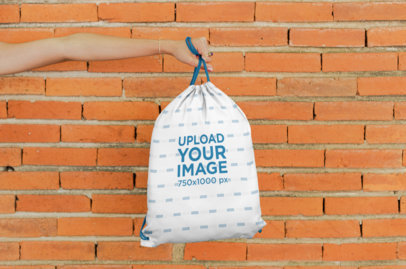 Mockup of a Hand Holding a Drawstring Bag Against a Brick Wall 27574