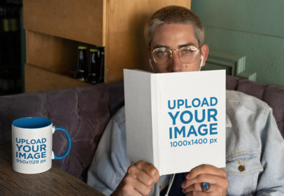 Mockup of a Blonde Man Looking From Behind a Book with an 11 oz Mug by His Side