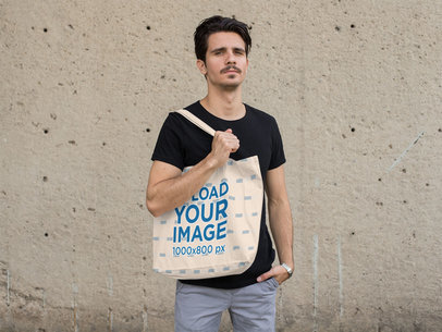 Mockup of a Man Holding a Tote Bag with a Concrete Wall in the Background 28855