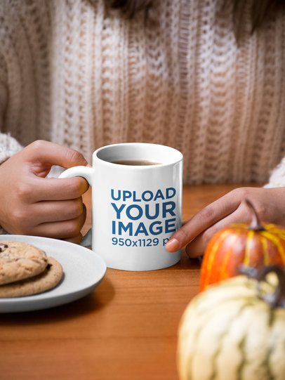 Mockup of a Woman Holding an 11 oz Mug with Cookies and Pumpkins at the Table