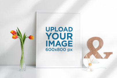 Mockup of a Picture Frame Standing Against a White Wall and Next to a Flower Vase 