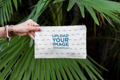 Mockup of a Hand Holding a Pouch Against Some Palm Leaves