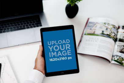 Mockup of a Man Holding an iPad Mini over His Work Desk 2112-el