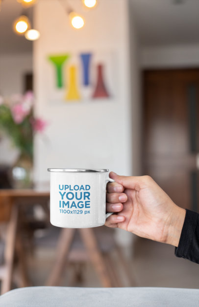 Mockup of a Hand Holding a 12 oz Silver Rim Enamel Mug at a Living Room 