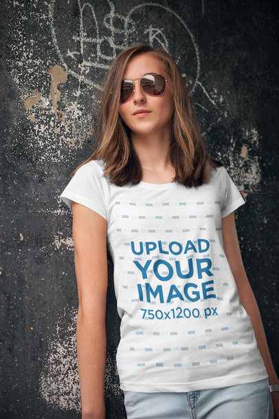 Mockup of a Young Woman Wearing a T-Shirt and Leaning Against an Urban Wall