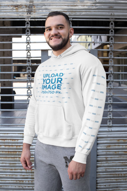 Mockup of a Man Wearing a Pullover Hoodie with a Customizable Sleeve Against a Metal Fence 