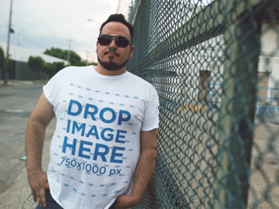 T-Shirt Mockup of a Guy Wearing Sunglasses Leaning Against a Fence