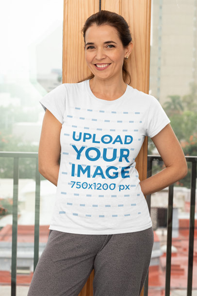 T-Shirt Mockup Featuring a Middle-Aged Woman Standing Against a Balcony Door 