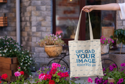 Mockup Featuring a Woman's Hand Holding a Tote Bag by Some Flower Pots 