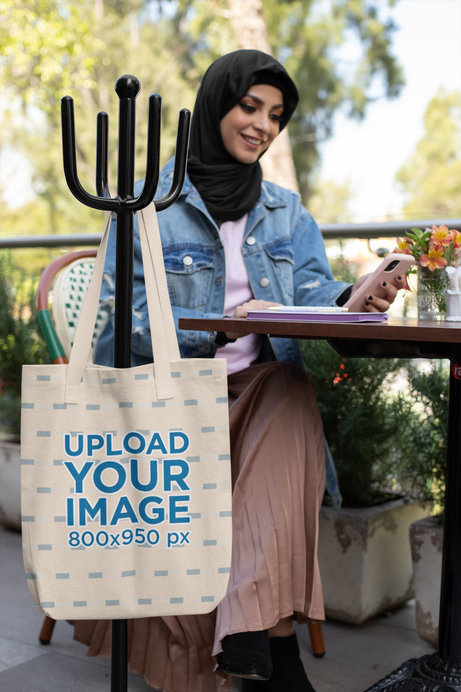 Tote Bag Mockup Featuring a Woman Wearing a Hijab and Using Her Phone at a Restaurant