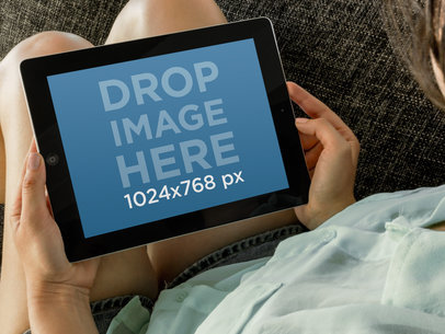 Mockup of a Woman Holding an iPad While Sitting on a Grey Fabric Couch  -