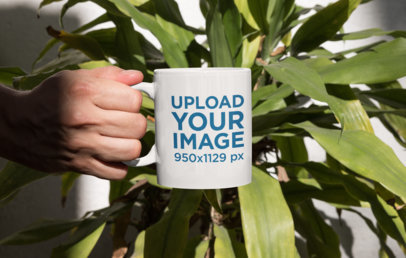 Mockup of a Person Holding an 11 oz Mug in Front of a Plant
