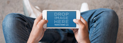 Mockup of Man Sitting on The Floor While Holding an iPhone in Landscape Position