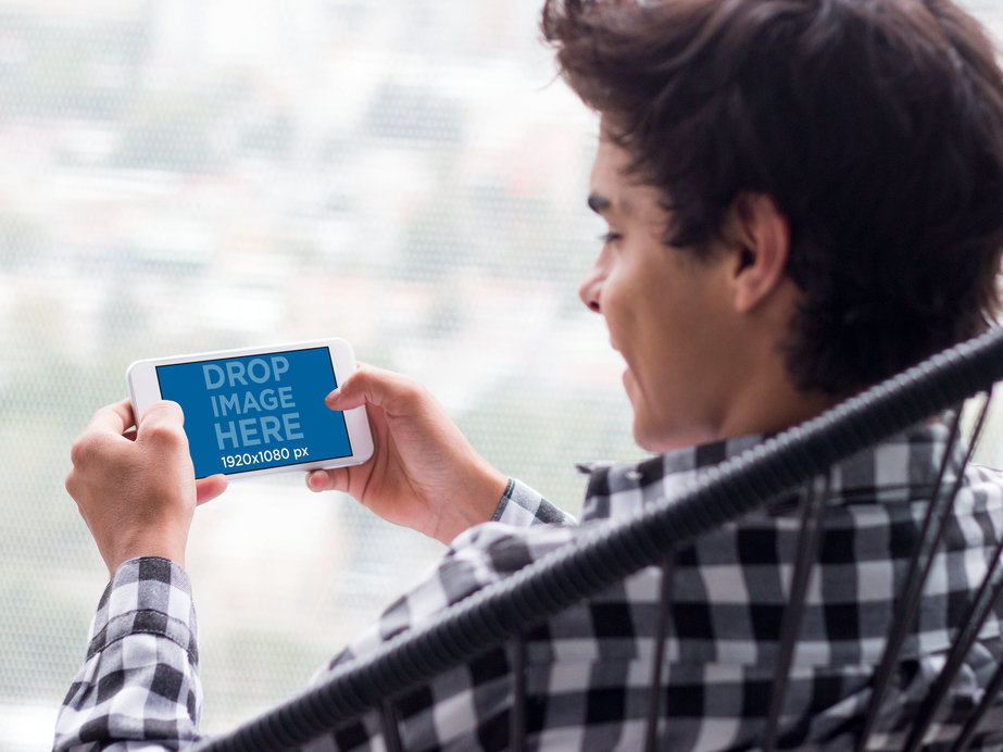 Mockup of a Young Man Staring at an iPhone in Landscape Position with Blurry Background