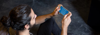 Mockup of a Hipster Man Sitting in the Floor Holding an iPhone in Landscape Position